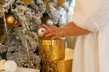 A woman is opening a gold box with a silver ball inside. The box is decorated with gold and silver ornaments, and the woman is wearing a white dress. The scene is set in a room with a Christmas tree.
