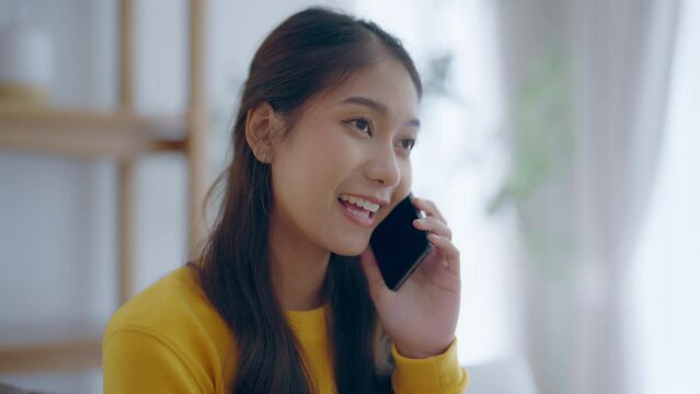Happy young asian woman speaking on mobile phone at home, Female teen answering call on cellphone while sitting in couch. Asia girl talking on smartphone