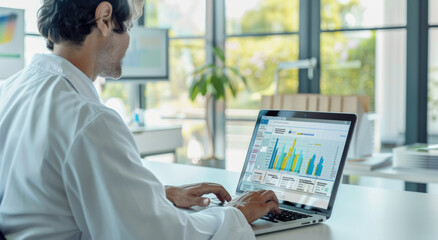 A doctor in a white coat is using a laptop with medical data on the screen while sitting at a desk.
