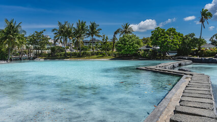 A winding concrete footpath crosses the turquoise water swimming pool. Cottages can be seen in the distance among the green tropical vegetation. Palm trees against a blue sky and clouds. Philippines. 