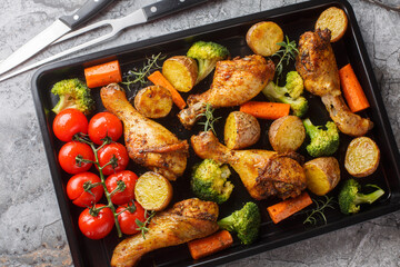 Tasty Baked ruddy chicken legs with potatoes, broccoli, tomato, carrot close-up on a baking sheet on the table. Horizontal top view from above