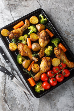 Baked chicken drumstick with broccoli, potatoes, tomatoes, and carrots close-up on a sheet pan on the table. Vertical top view from above