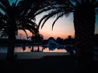 Fototapeten Soleil couchant sur une grande piscine avec les silhouettes des palmiers. Crépuscule sur un camping de vacances. Ambiance détente. Se reposer et admirer le paysage de la piscine et des palmiers.  © david