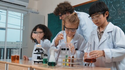 Caucasian smart teacher support excited curious student pour colored liquid in beaker at laboratory. Creative children concentrate to do experiment and enjoy inspecting biochemical sample. Pedagogy.