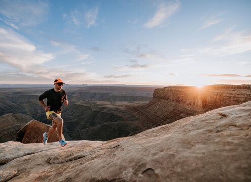 Male trail runner jogs along mesa edge in Utah desert, sunset.