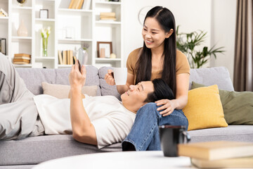 Happy Asian couple relaxing at home, man lying on couch using smartphone with smiling woman beside him holding a coffee mug.