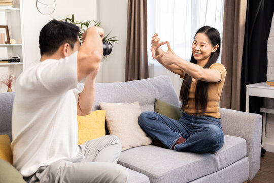 A young man takes a photo of a woman sitting comfortably on a sofa in a cozy living room setting.