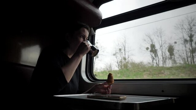 A girl eats a croissant and drinks coffee or tea while sitting at a table in a train compartment and looks out the window at nature. A woman travels by rail.