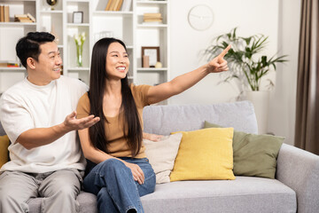 Asian Couple Pointing sitting on the sofa engaging in conversation, with one partner pointing to something unseen and discussing it animatedly.