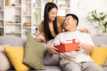 Asian couple enjoying a surprise gift moment on sofa at home, conveying happiness, celebration, and loving relationship