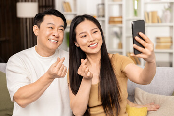 Happy Asian Couple Taking Selfie At Home. Smiling Man And Woman Posing For A Cheerful Mobile Photo In A Stylish Living Room Setting, Embodying Happiness And Modern Lifestyle.