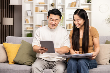 Couple sitting together on a sofa, engaged in paperwork and digital tablet in a cozy living room.
