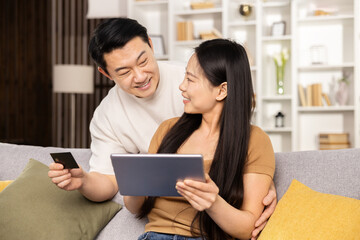 Asian couple shopping online together on tablet, man holding credit card, happy, cozy home interior