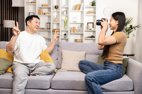 A cheerful man poses with a peace sign while a woman focuses on taking his photograph with a DSLR camera in a cozy living room.