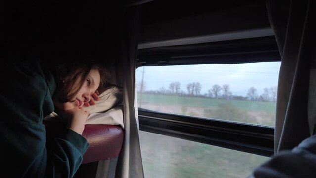 Teenage girl lies on top bunk of train compartment and thoughtfully looks into distance through open window. Travel, train ride. School girl rests lying on a bunk on a train.