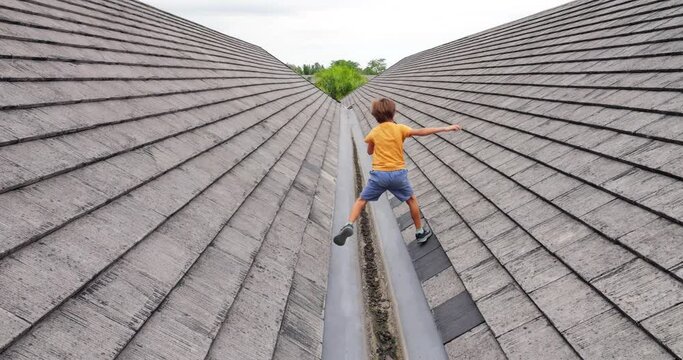 Young boy playfully jumps over gutter between two slopes of roof, captured in slow motion from behind. His nimble and energetic movements showcase his adventurous spirit