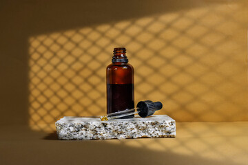 Blank amber glass essential oil bottle with pipette on brown  background with twist mesh window shadow. Skin care concept with natural cosmetics
