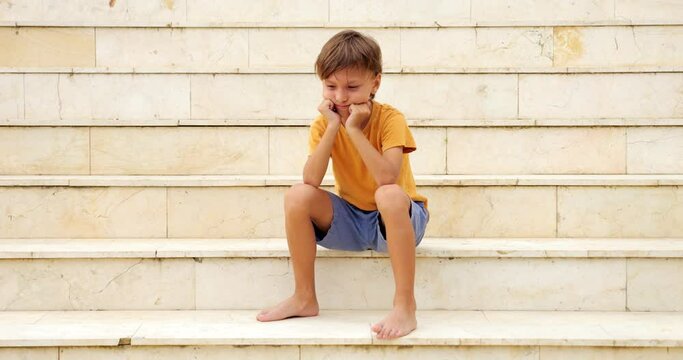 Young boy appears upset as he puffs his cheeks and rests his head on his fists. He raises his head slightly and gazes unhappily into camera, seemingly seeking words of encouragement or positivity.