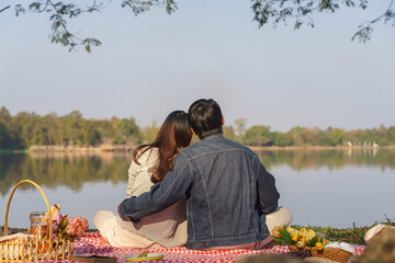 Young couple in love sitting on blanket with picnic basket and embracing to watching lake view - 784339097