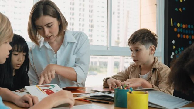 Professional caucasian teacher telling story to diverse student while sitting at table with storybook and colored book. Smart learner listening story while colored picture from instructor. Erudition.