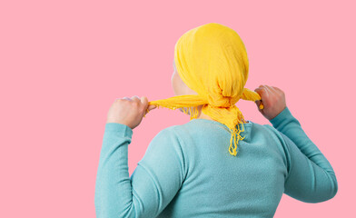 Young woman from behind putting a yellow scarf on her head because she has cancer on a pink background