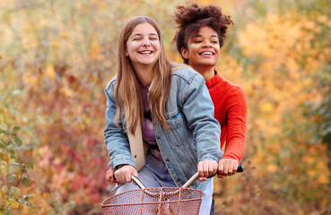 Two young happy diverse female best friends riding bicycle together in autumn forest