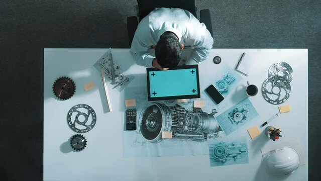 Top view of engineer looking at phone while working on tablet with green screen at meeting table with turbine engine or jet engine sketch design and various metal gear scatter around. Alimentation.