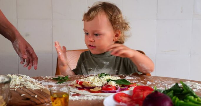 Small kid watches as mom sprinkle grated cheese on top of homemade pizza, he's the head chef. Amusing scene, young child helps his mother on the kitchen, messy table with flour all around.