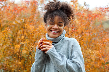 Young happy smiling mixed-race woman with coffee cup in autumn nature, pleased african american female with curly hair in knitted sweater
