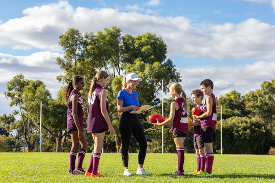 smiling female coach holding clipboard addressing schoolchildren in sports uniforms