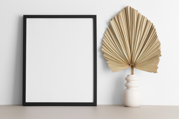 Black frame mockup with a palm leaf decoration on the beige table.