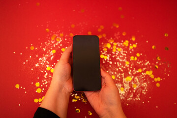 Beautiful female hands hold a phone over a red background with silver sparkles and gold round...