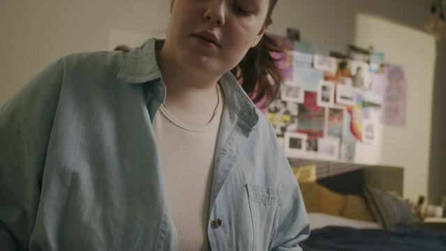 Waist up of Caucasian female high school or college student girl taking notes on sheet of paper while studying in cozy dorm room with posters hanging on wall