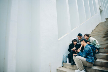 Four Asian university students sit on the campus steps discussing their coursework, simultaneously...