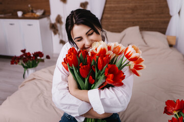 A woman poses on bed, surrounded by vibrant fresh bouquets bunch of red tulips placed around the room, domestic atmosphere. Spring International Women's Day, 8th of March. Holiday, festive 
