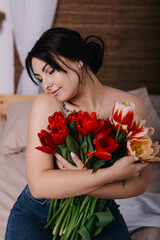 A woman poses on bed, surrounded by vibrant fresh bouquets bunch of red tulips placed around the room, domestic atmosphere. Spring International Women's Day, 8th of March. Holiday, festive 
