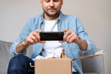 A man in a denim shirt unpacks a parcel and takes a photo of his purchase, internet order. Person...