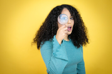 African american woman wearing casual sweater over yellow background surprised looking through a magnifying glass