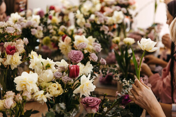 Hands delicately holding white tulips amidst a vibrant assortment of roses and daffodils during a bustling floral arrangement workshop.