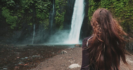 A woman stands at the base of a majestic Bali waterfall, surrounded by lush greenery. Water...