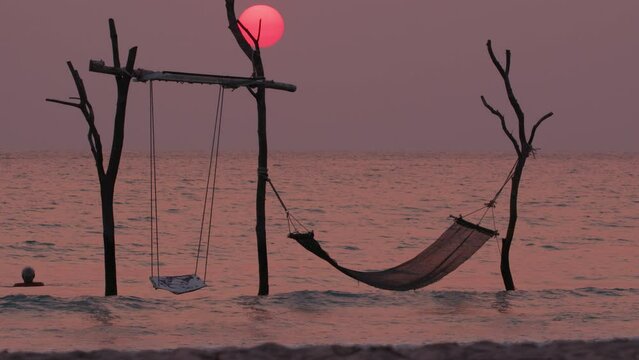 Sea sunset landscape with swing and hammock on background of red sun in sky. Concept of tourism and beach vacation at resort.