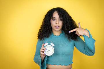 African american woman wearing casual sweater over yellow background surprised holding and pointing a clock