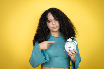 African american woman wearing casual sweater over yellow background serious holding and pointing a clock