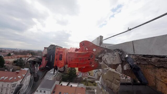 FPV drone view of A demolition excavator with a hydraulic hammer attachment powerfully breaking through a concrete wall, amid the rubble of a building being deconstructed.
