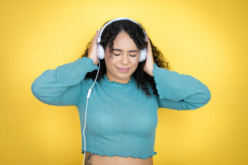 African american woman wearing casual sweater over yellow background listening to music using headphones over yellow background singing and screaming