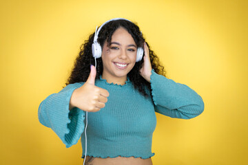 African american woman wearing casual sweater over yellow background using headphones success sign doing positive gesture with hand, thumb up smiling and happy. cheerful expression