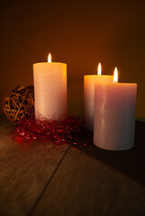Three candles are lit on a table with a red ball in the background. The candles are white and yellow, and the red ball is made of wood. The scene has a warm and cozy atmosphere