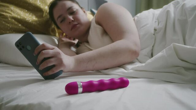 Young Caucasian sleepy woman waking up in bed early in morning, checking up time on her cellphone and falling asleep again. Purple adult toy lying beside her