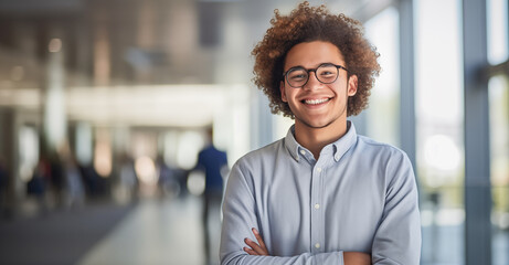 A cheerful young man with curly hair and glasses standing confidently in a bright office hallway, arms crossed, exuding a welcoming, professional vibe.