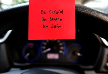 Red sticky note with text written BE CAREFUL BE AWARE BE SAFE , on the car steering wheel , to...